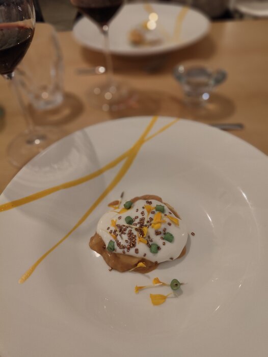 Väldekorerad dessert på tallrik, glas rödvin, elegant middagsmiljö, blomblads- och såsgarnityr.