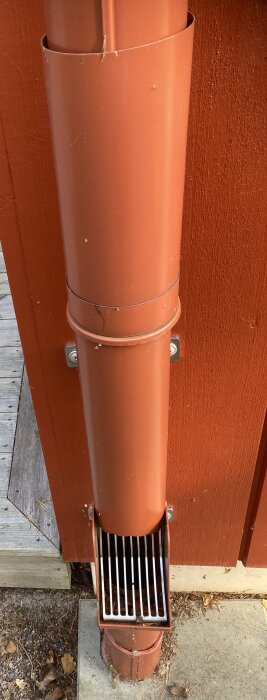 Röd stupränna på en trävägg, metallgaller nedtill, omgiven av trä och betongytan.