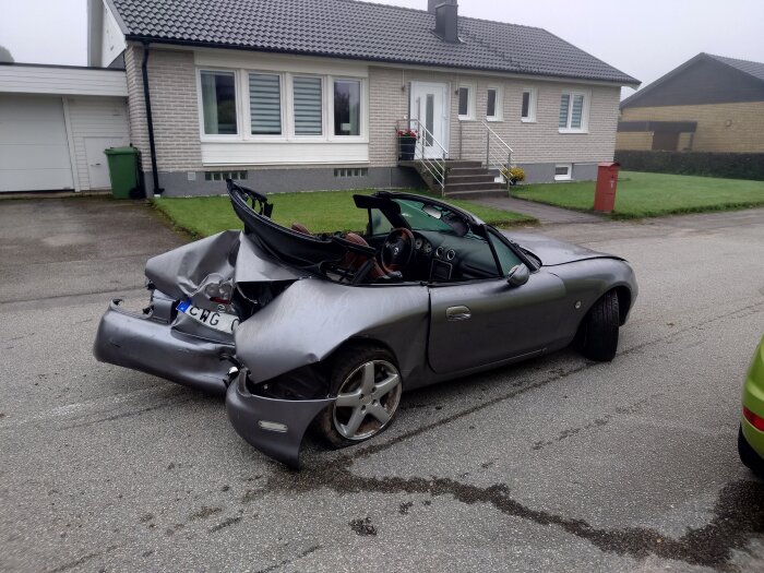 Grå cabriolet med omfattande skador efter en olycka, parkerad framför ett hus.