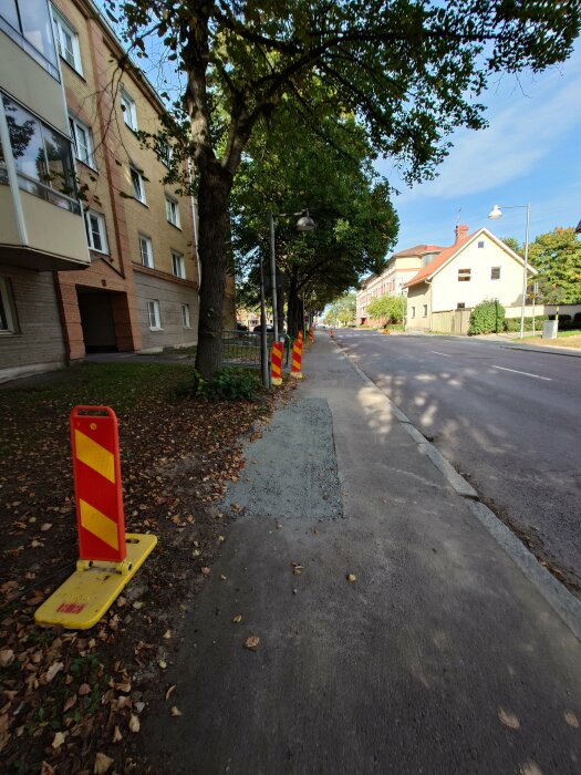 Stadsgata med trafikkoner, trottoar, byggnader, träd och blå himmel på en solig dag.