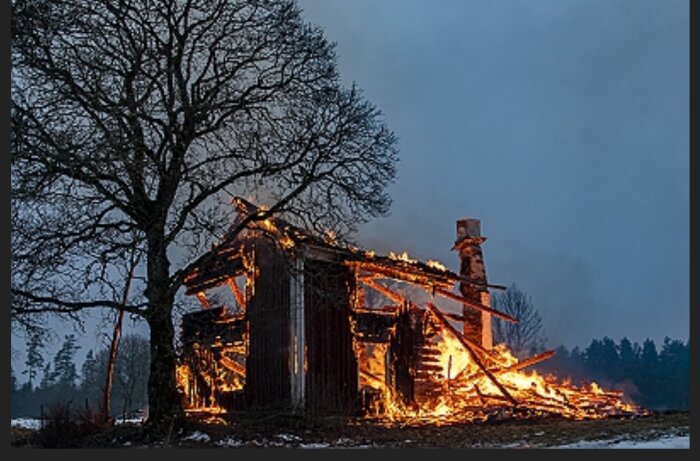 Ett trähus brinner i skymningen, omgivet av träd, med synliga lågor och rök.