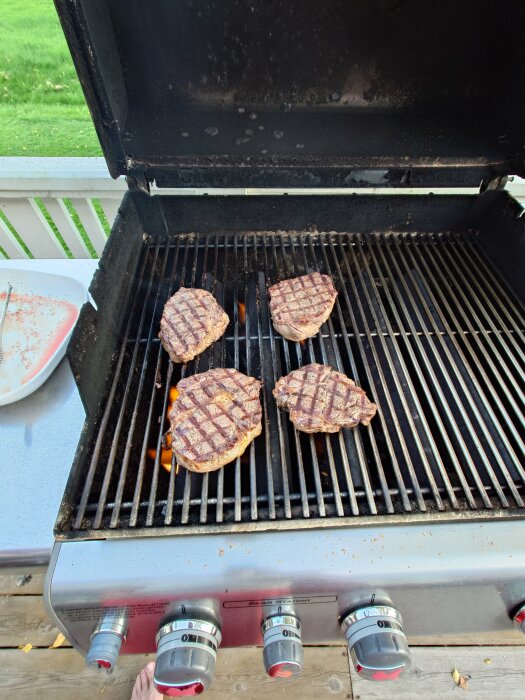 Fyra biffar grillas på en gasolgrill på en altan, utomhusmiljö, grillverktyg och barfota person synliga.