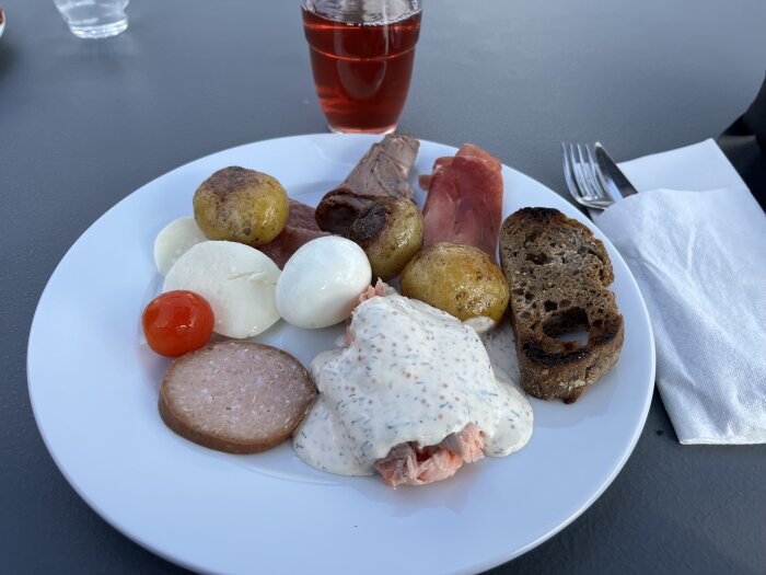 En tallrik med blandad mat: potatisar, mozzarella, kött, lax, tomat, rågbröd, skinka och dryck.