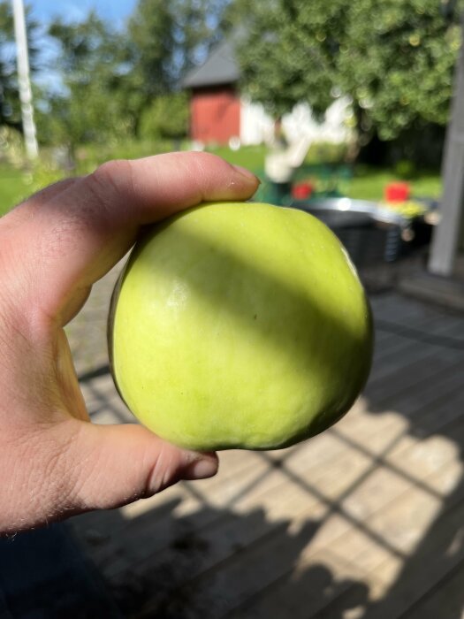 Hand håller ett grönt äpple, oskarp trädgård i bakgrunden, solljus, träterrass, sommarmiljö.