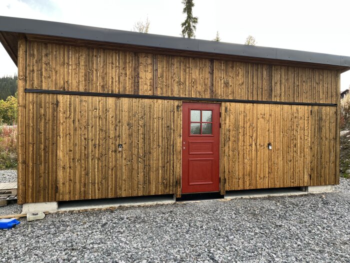 Träskjul med röd dörr, svart tak, grusmark framför, detaljer i metall, ljus himmel, delvis skymd naturbakgrund.