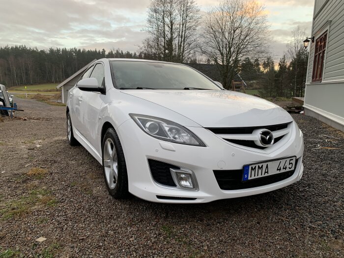 En vit Mazda parkerad på en grusuppfart, framför ett hus, med skymmande träd i bakgrunden.