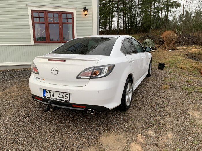 Vit Mazda6 parkerad vid en byggnad, grusgång, landsbygdsmiljö, dagtid.