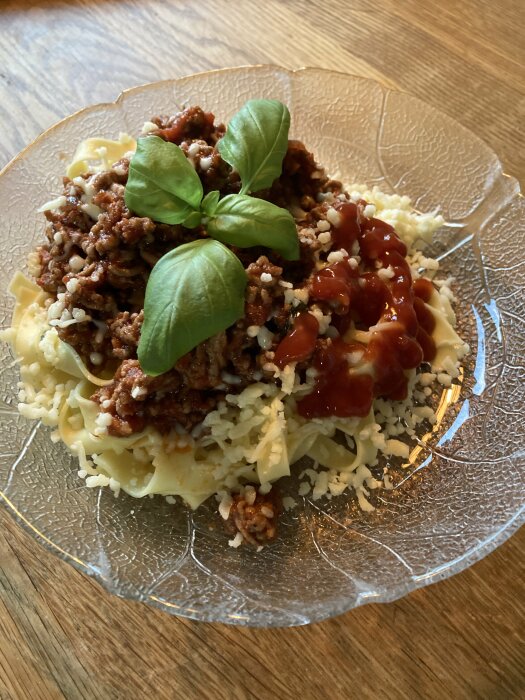 Pasta med köttfärssås, riven ost, ketchup och färsk basilika på ett glasfat.