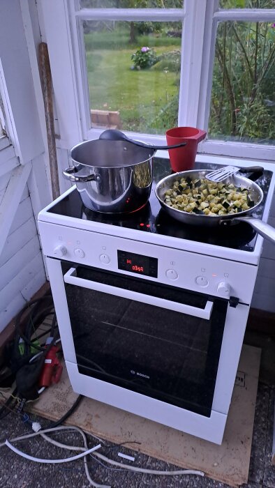 Spis med kokande kruka och stekpanna, fönsterutsikt mot trädgård, oorganiserat utomhuskök, kvällsljus.