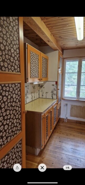 Rustik kök, träbjälkar, blommiga tapeter, orange skåpsluckor, fönster med utsikt mot grönska.