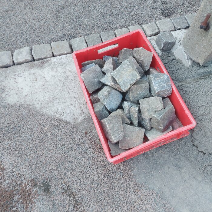Röd låda fylld med grå stenplattor; gatsten läggs; stenläggning pågår.