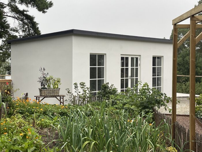Ett vitmålat hus med fönster bakom en blomstrande trädgård och en korg på en bänk.