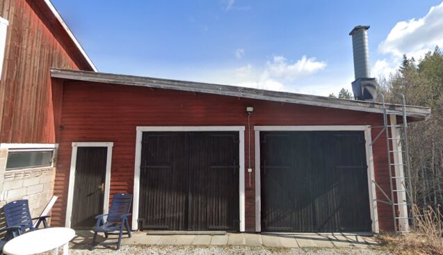 Röd lada med två garageportar och en vit dörr, stolar framför, dagtid, delvis molnig himmel.