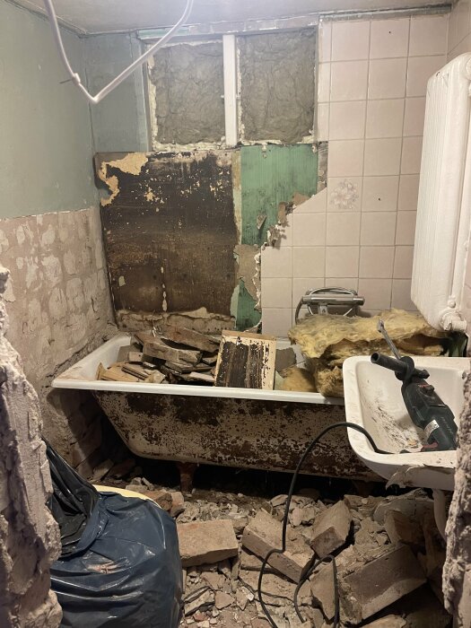 Förfallen badrum; trasiga väggar, skräp, gammal badkar, renovering pågår med synlig borr.