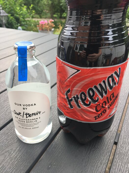 Vodkaflaska och cola på träbord utomhus, speglade grönska, zero sugar, dagtid.