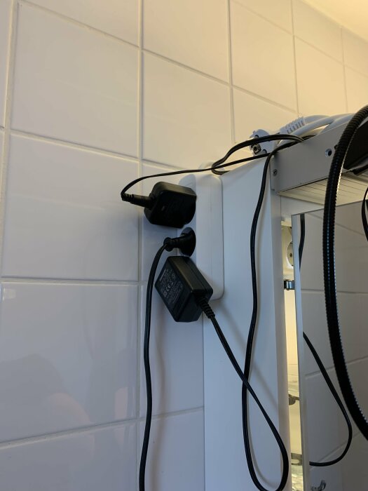 Vit kakelvägg, eluttag, två elektriska apparater inkopplade, sladdar, vit möbelkant.