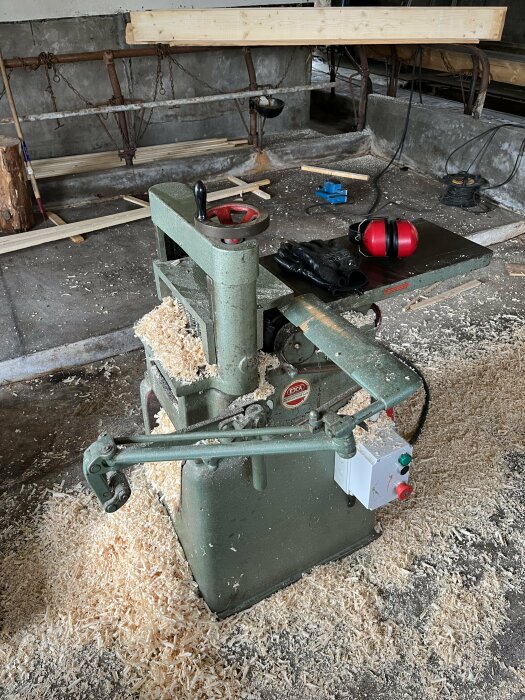 Verkstad med träspån, stationär träbearbetningsmaskin, säkerhetsutrustning, verktyg, arbetsbänk och oordning.