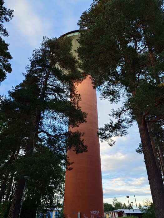 Hög vattentorn, träd runtomkring, blå himmel med moln, gatlyktor, graffiti på tornets botten.