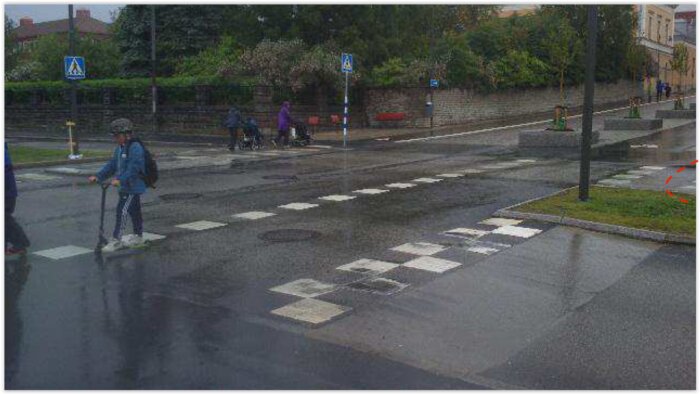 Regnig gata med övergångsställe, personer, blå skyltar, våt asfalt, gröna växter och en sparkcykel.