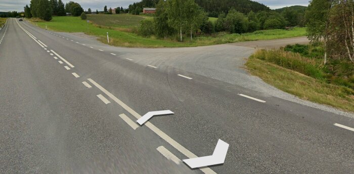 Lantlig vägkorsning med vägmarkeringar och grönområden, delvis molnig himmel, ingen trafik observerbar.