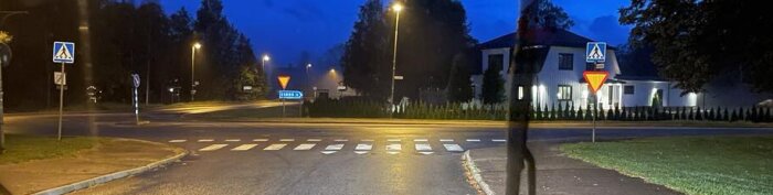 Kvällsbild av en öde vägkorsning med övergångsställe, trafikskyltar och belysning nära byggnader.