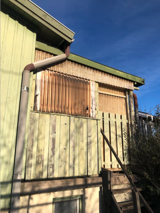 Ett grönt trähus med rostig plåt och en trappa under en klarblå himmel.