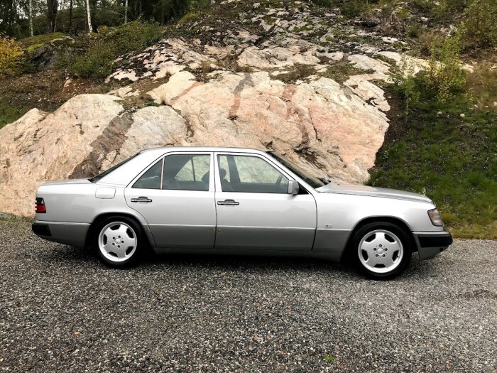 Grå sedanbil parkerad framför klippiga backar och grönska. Klassisk design, rena linjer, inga personer synliga.