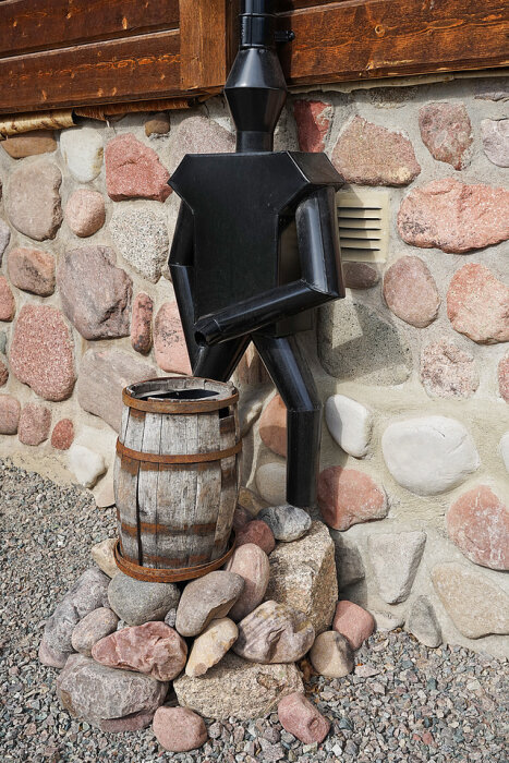 Stiliserad svart skorstensfigur med borste, stenmur, träfönsterlucka, ekfat, grus. Dekorativ, utomhus.