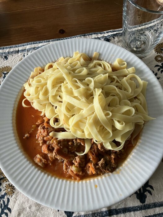 Tallrik med pasta och köttfärssås, vatten i glas, bordduk, måltidssituation.