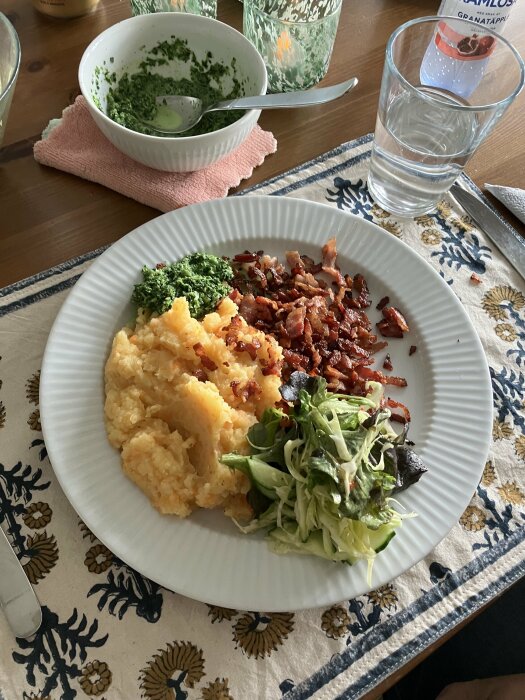 Middagstallrik med potatismos, knaperstekt bacon, grönsallad, pesto, vattenglas och öppnad matdryck på duken.