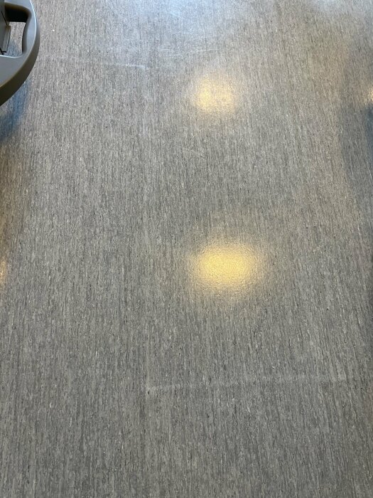 Texturerat grått golv med ljusa reflektioner, möjligen från överliggande belysning. En del av en möbel syns.