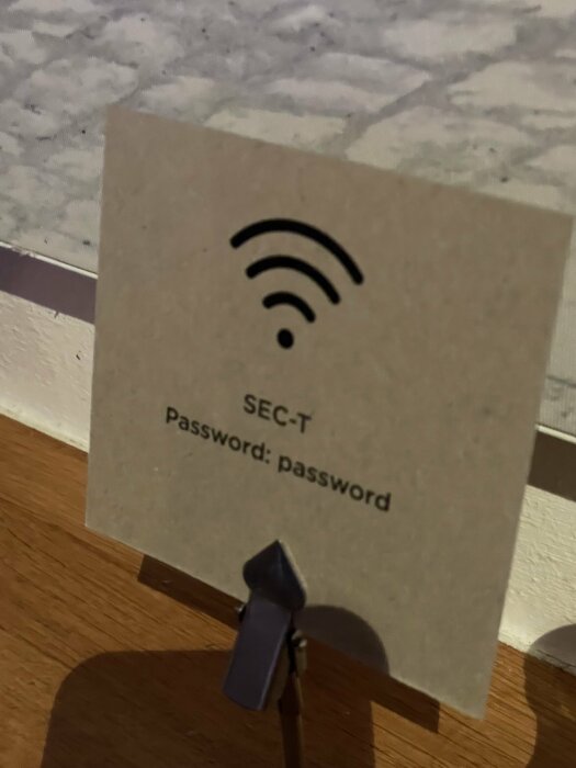 WiFi-symbol, text "SEC-T" och "Password: password" på papper, står med träklämma och skugga på bordet.