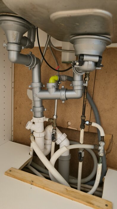 Komplext rörsystem under diskbänk med avloppsrör, vattenledningar och avstängningsventil, synligt i köksskåp.