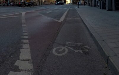 Cykelbana på gata vid gryning/skymning, vit cykelmarkering, tom väg, trottoarkant och gatubelysning.