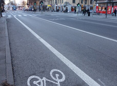 Stadsgata med cykelbanemarkering, fordon, fotgängare, och stadsmiljö i bakgrunden. Klar dag och asfalterade vägar.