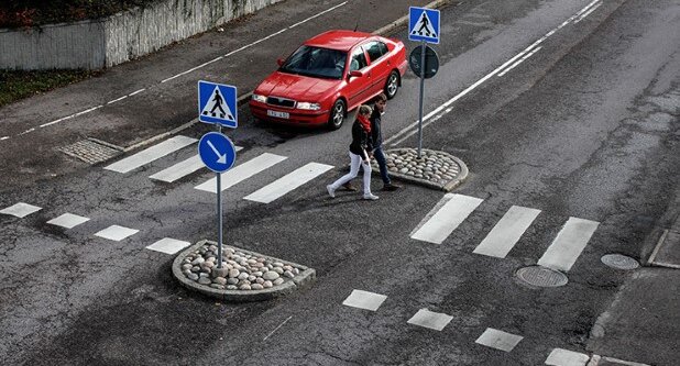 Två fotgängare korsar vägen vid ett övergångsställe, röd bil väntar, trafikskyltar synliga.