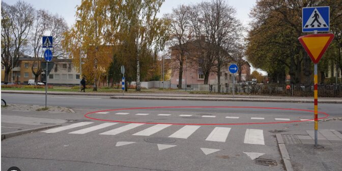 Stadsvy med övergångsställe, trafikskyltar, trädkantad gata, och en ensam fotgängare i bakgrunden.