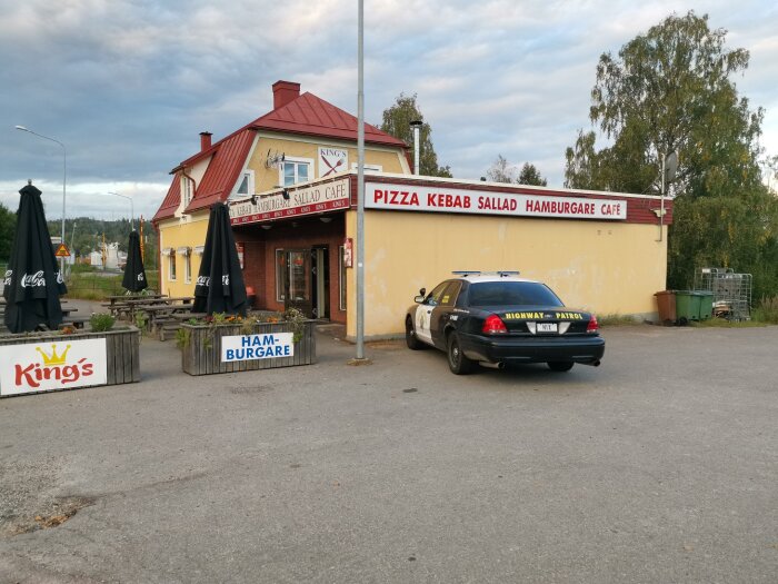 Ett gult hus med skyltar för pizza, kebab, sallad. Utomhusbord med svarta parasoller. Polisbil parkerad bredvid.
