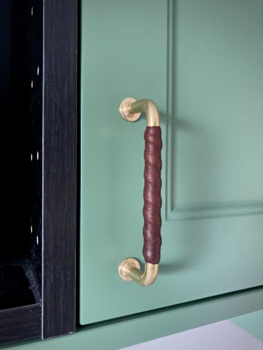 Läderhandtag med guldfärgade beslag på en grön dörr, bredvid svart dörrkarm.