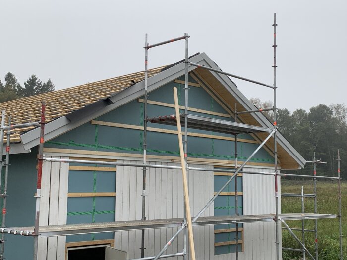 Hus under konstruktion med ställningar, träspikremsa och gröna väderskyddsskivor, molnig himmel.