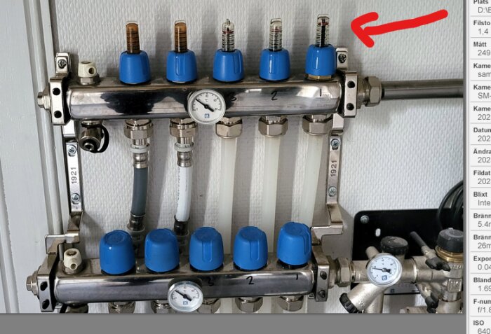 Värmesystemets distributionsblock med rör, ventiler, mätare och blå rattar för justering. Röd pil pekar på objekt.