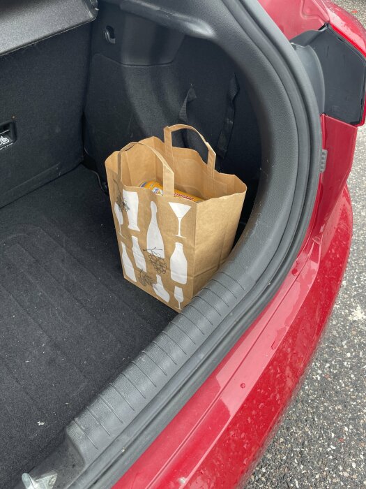 En papperskasse med varor i ett öppet bilbagage, röd bil, grusig mark, synbara små stänk på bil.