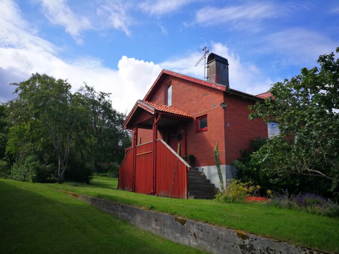 Rött tegelhus med veranda, grönt gräs, träd, blå himmel och vita moln.