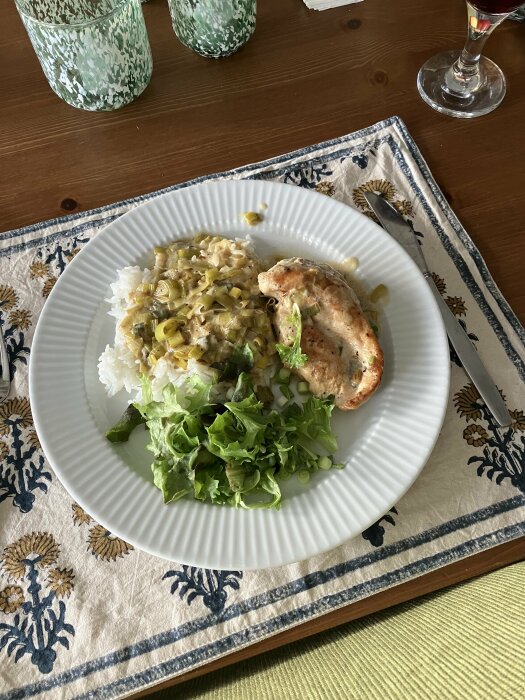 Tallrik med ris, grön currysås, kyckling och sallad; vattenglas och vin; mönstrad löpare på träbord.