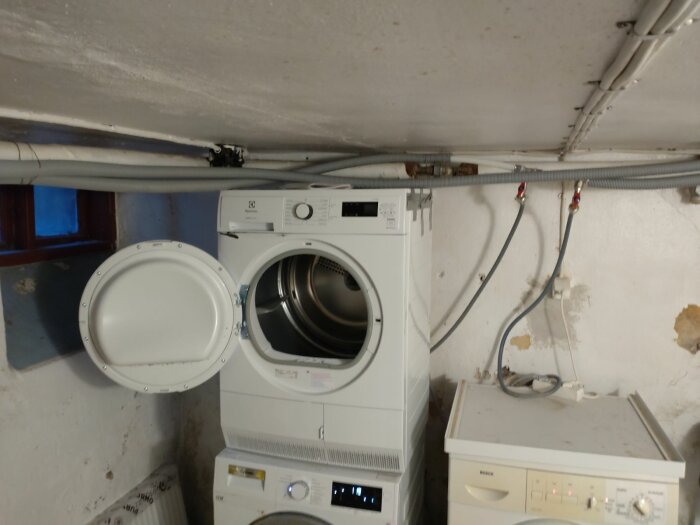 Tvättmaskin och torktumlare i ouppfräschat källarrum med synliga rör och sladdar.
