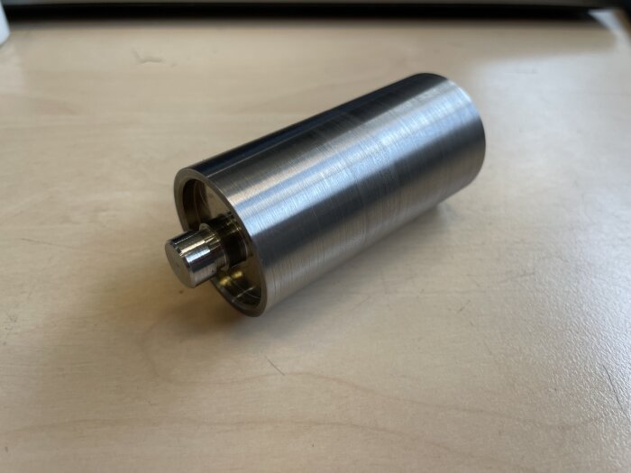 Metallisk cylinder med axel på ljus bordsyta, möjligen en mekanisk komponent eller del.