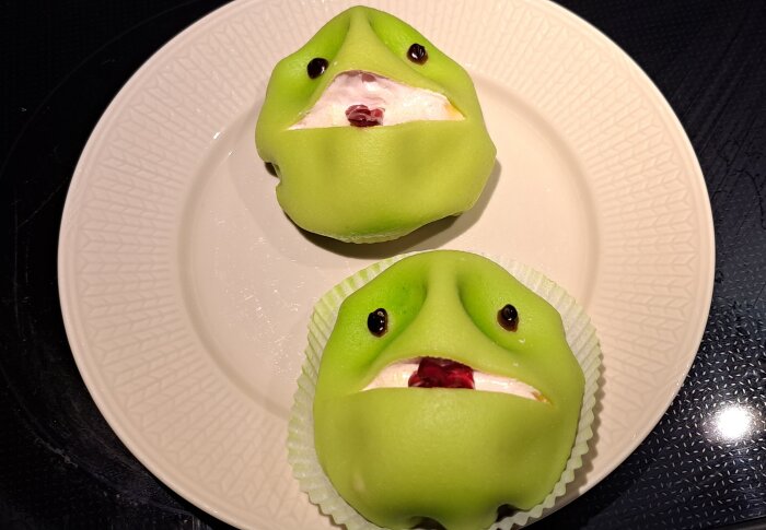 Två gröna bakverk med ansiktsuttryck, liknar hippos, på tallrik. Kreativt, humoristiskt, dessertkonst.