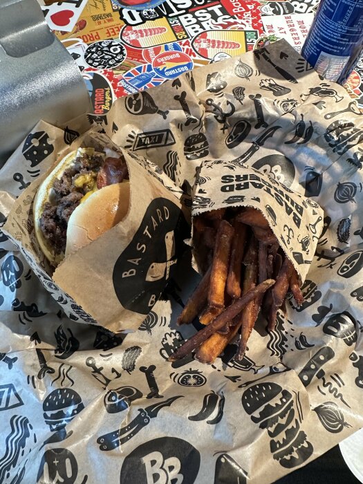 Hamburgare och sötpotatisfries på tryckt papper, snabbmatsmiljö, dryck i bakgrunden, måltid redo att ätas.