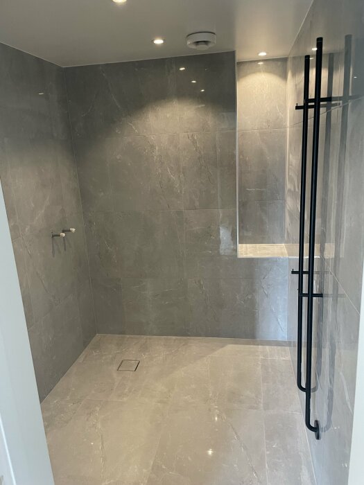 Modernt badrum, grå marmorplattor, glasduschvägg, svarta detaljer, takdusch, infällda spotlights, minimalistisk design.