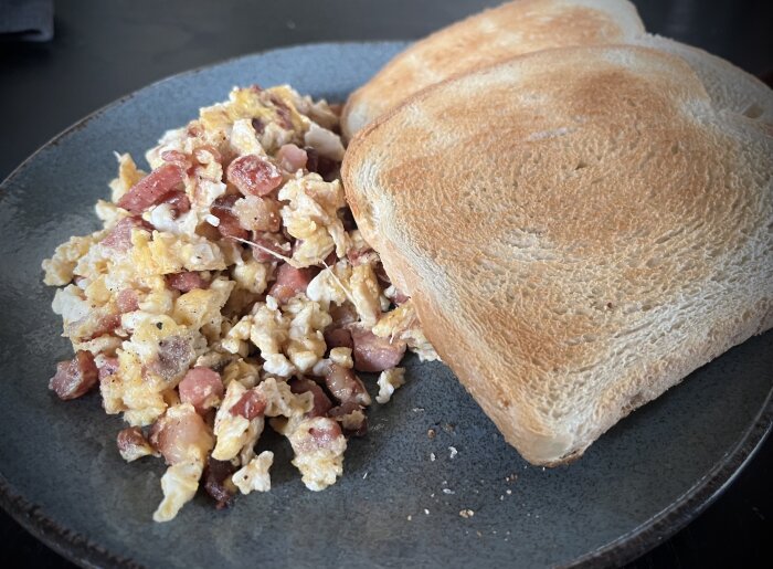 Stekt ägg med tärnad korv och brödskivor på en mörk tallrik. Frukostmåltid. Enkel presentation.